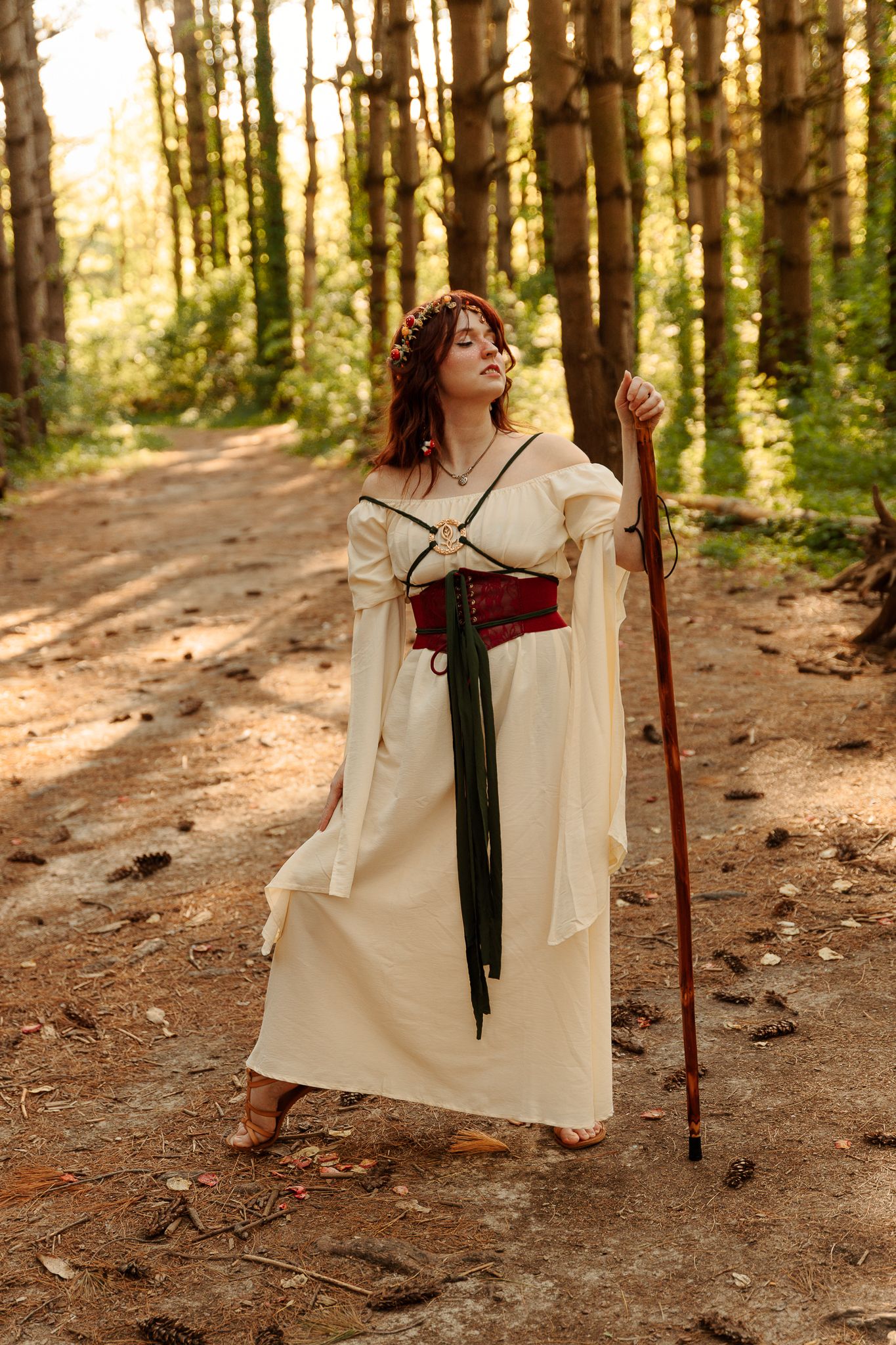 Woman in medieval fantasy costume with staff and corset belt standing barefoot on forest path in sunlight.