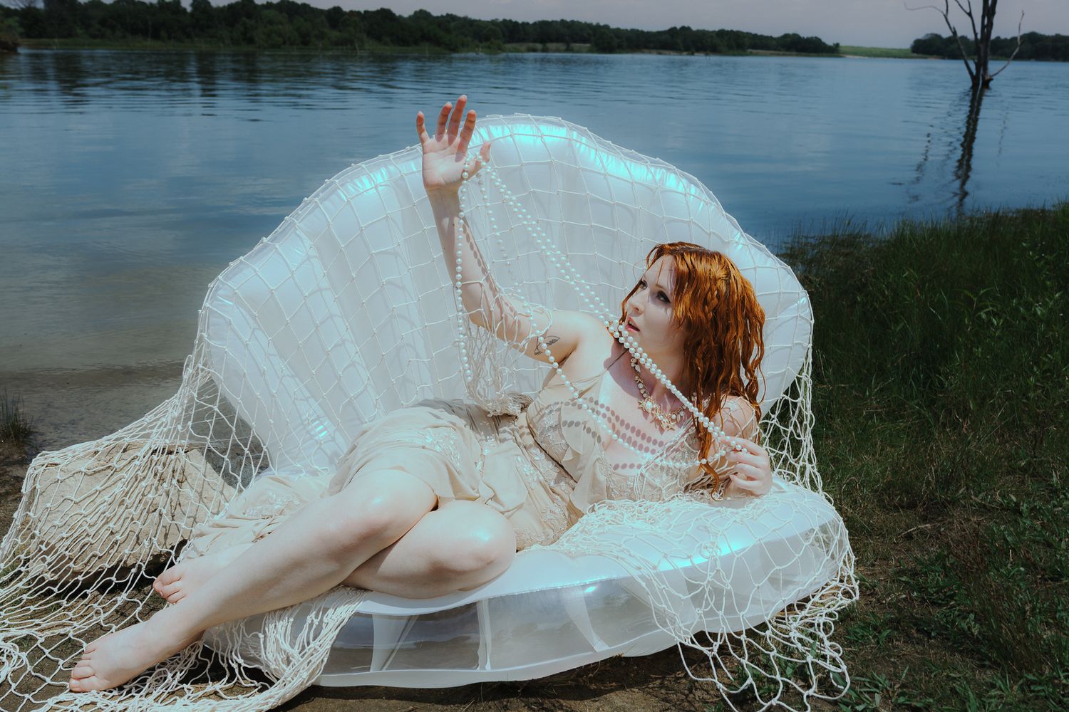 A red-haired woman reclines gracefully under a glowing net veil by a serene lake at dusk.