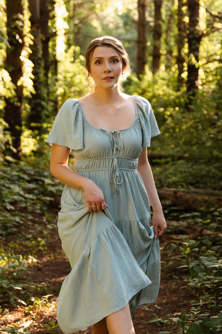 Person in flowing light blue dress walks through sunlit forest setting in romantic regency-style photoshoot.