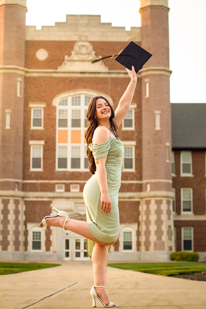 A graduate poses in a sage green dress outside a brick university building at sunset striking multiple poses.