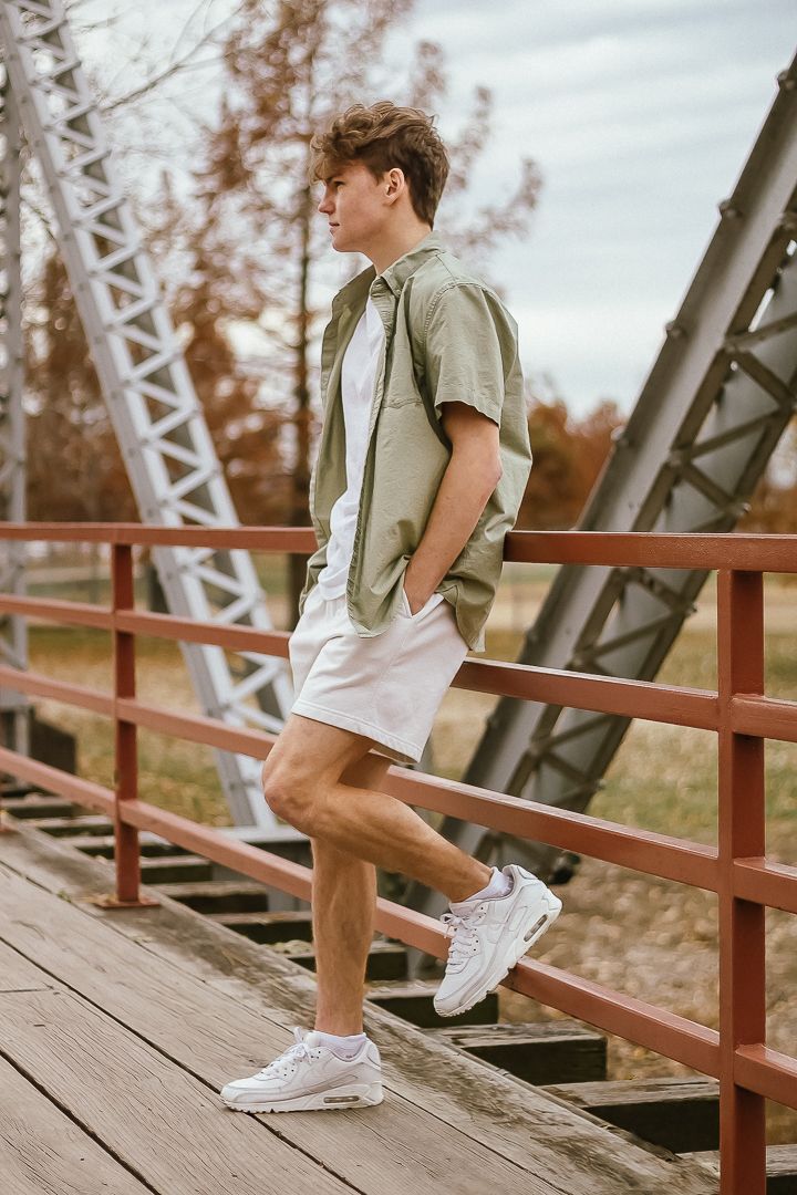 A person wearing white shorts and sneakers sits casually on a wooden bridge railing in fall scenery.