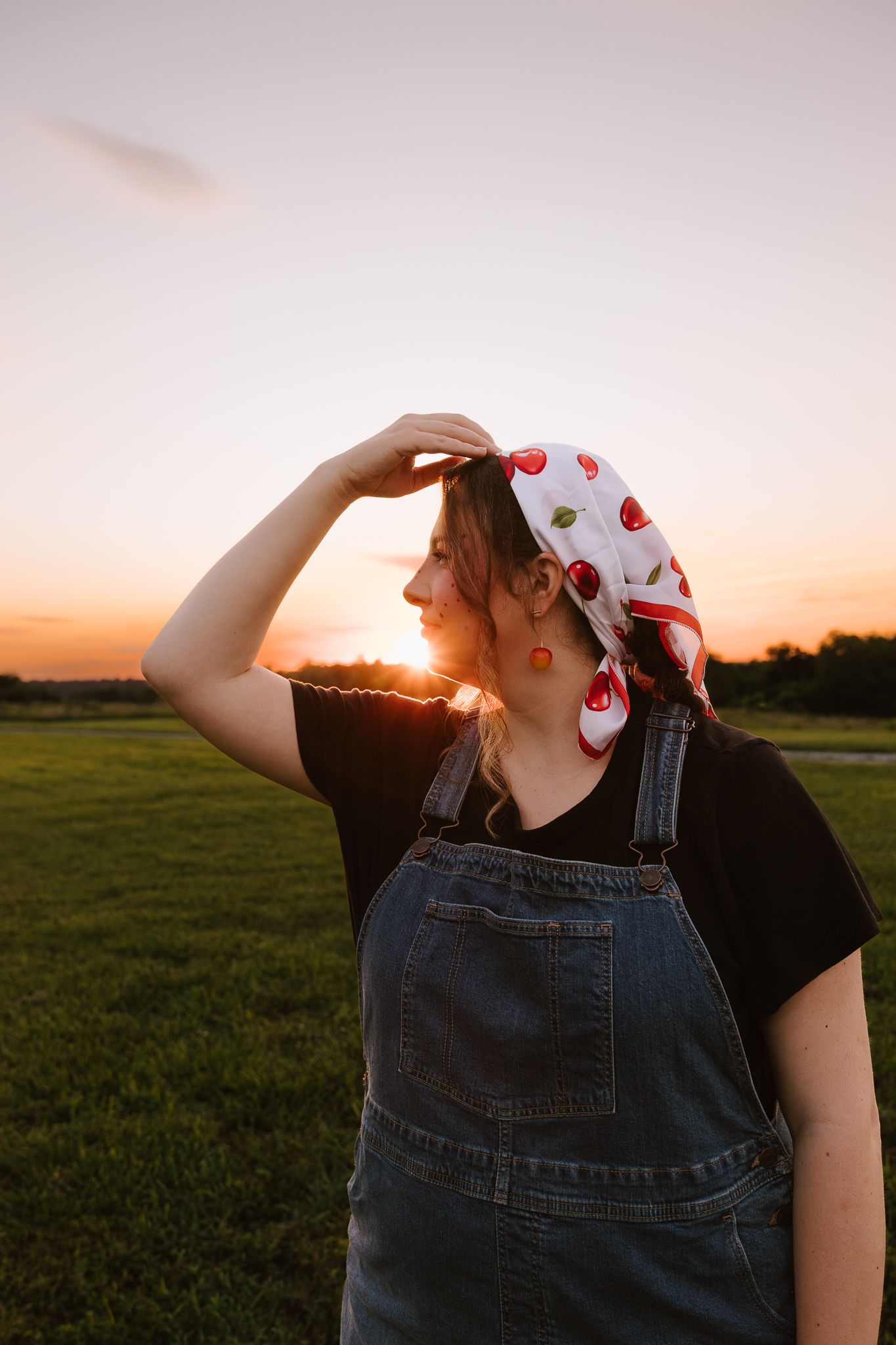 Young woman in denim overalls and cherry print bandana shielding eyes from golden sunset in green field.