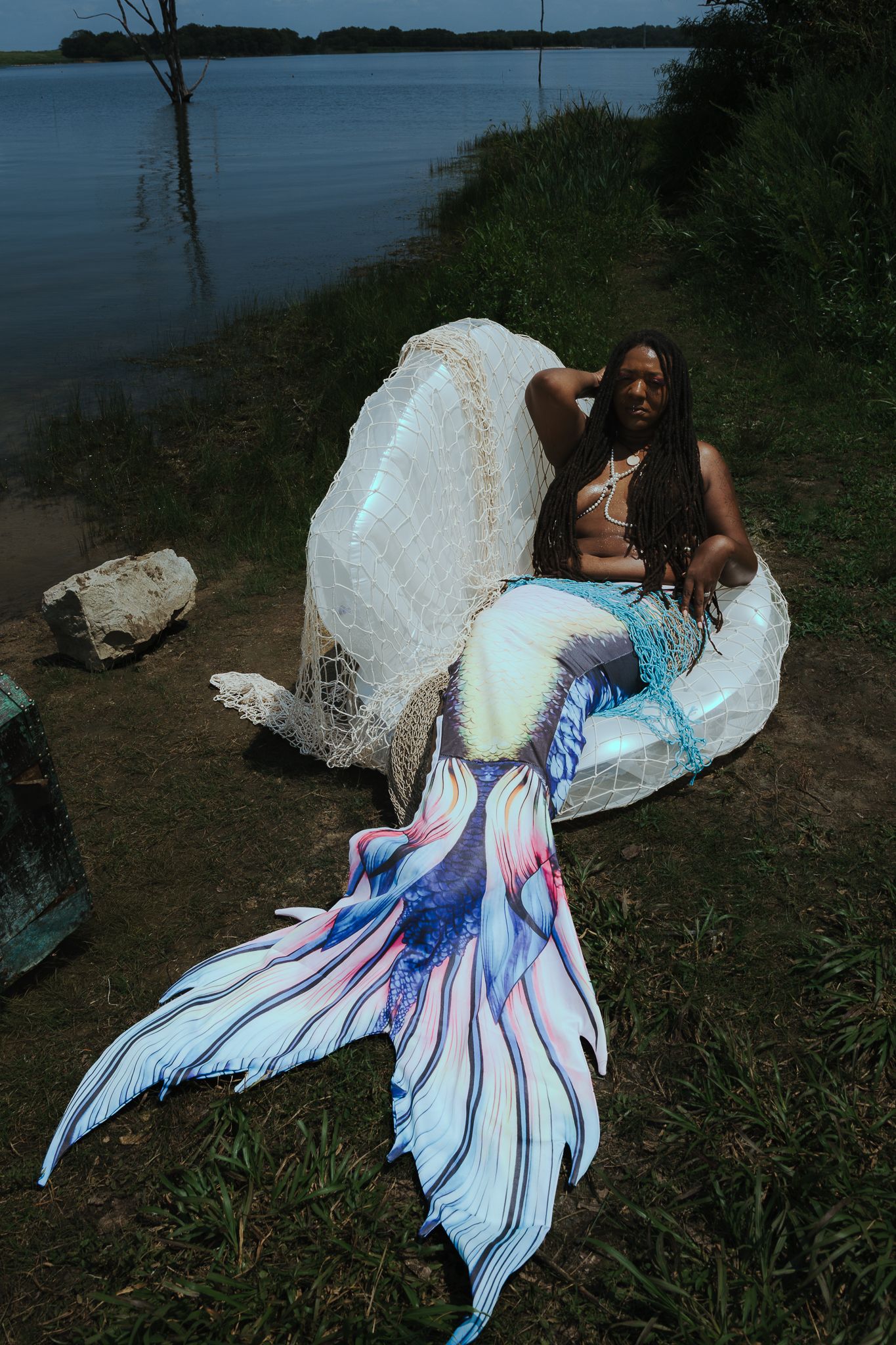 A Black woman in a colorful mermaid costume with tail poses by a lake shore surrounded by greenery.