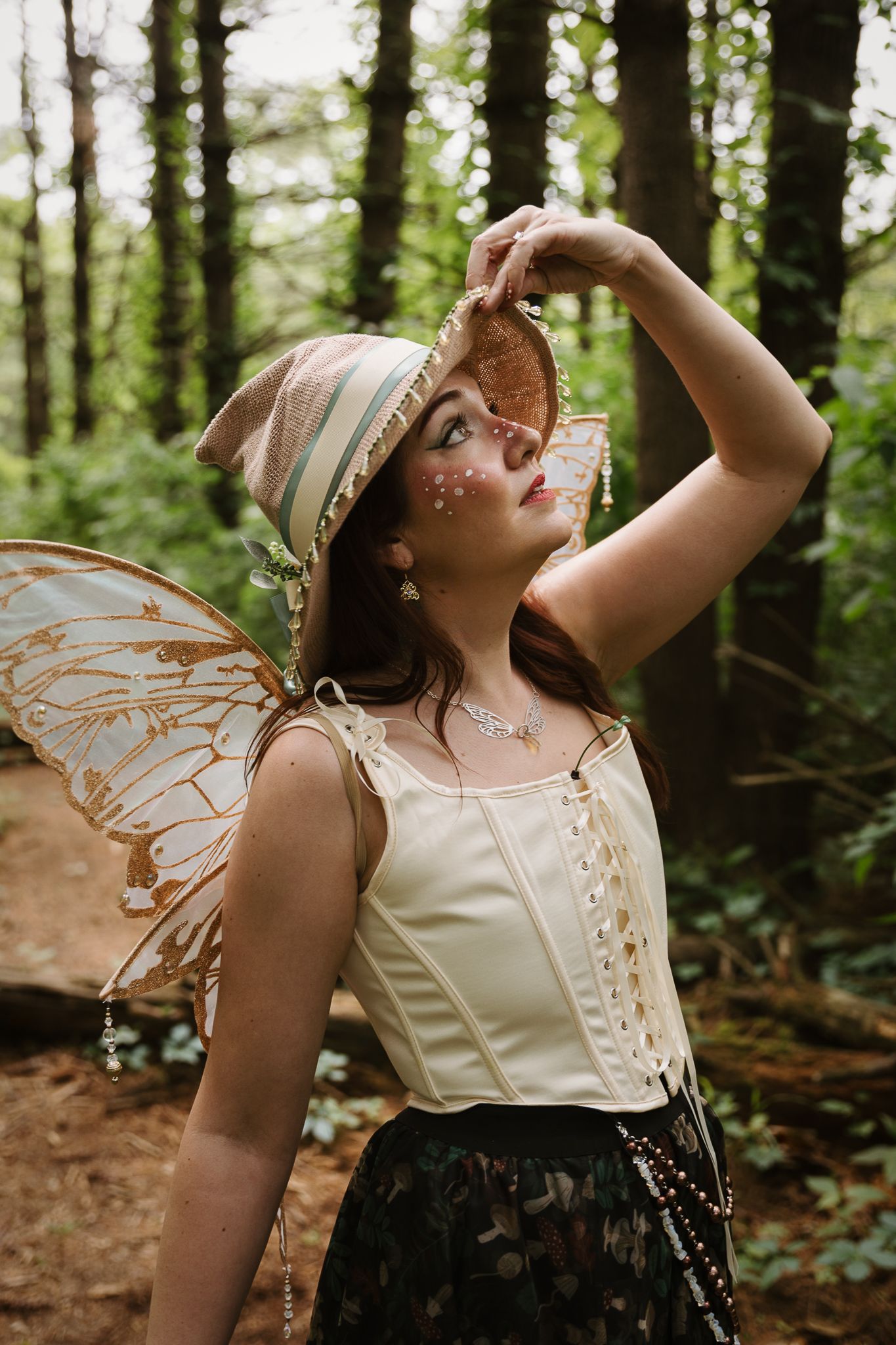 Woman in fairy costume with butterfly wings, corset, and floral hat poses in a lush green forest setting.