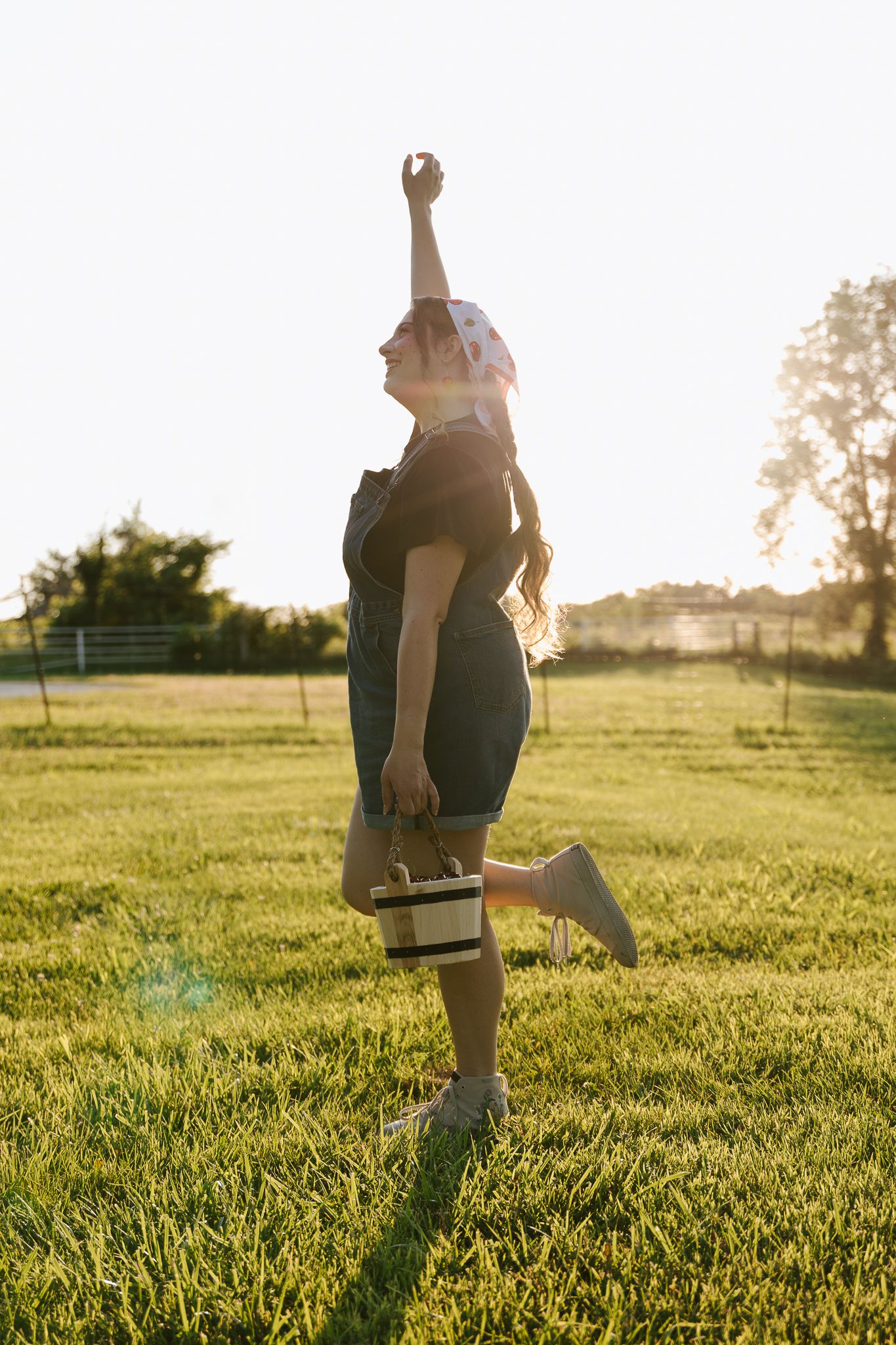 Woman in denim overalls celebrating with raised fist in sunny green field with lens flare.