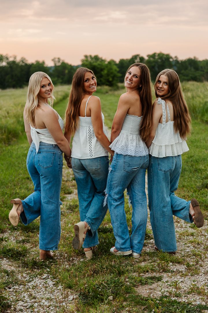 Friends in matching denim and white tops pose playfully together in a grassy field at sunset.