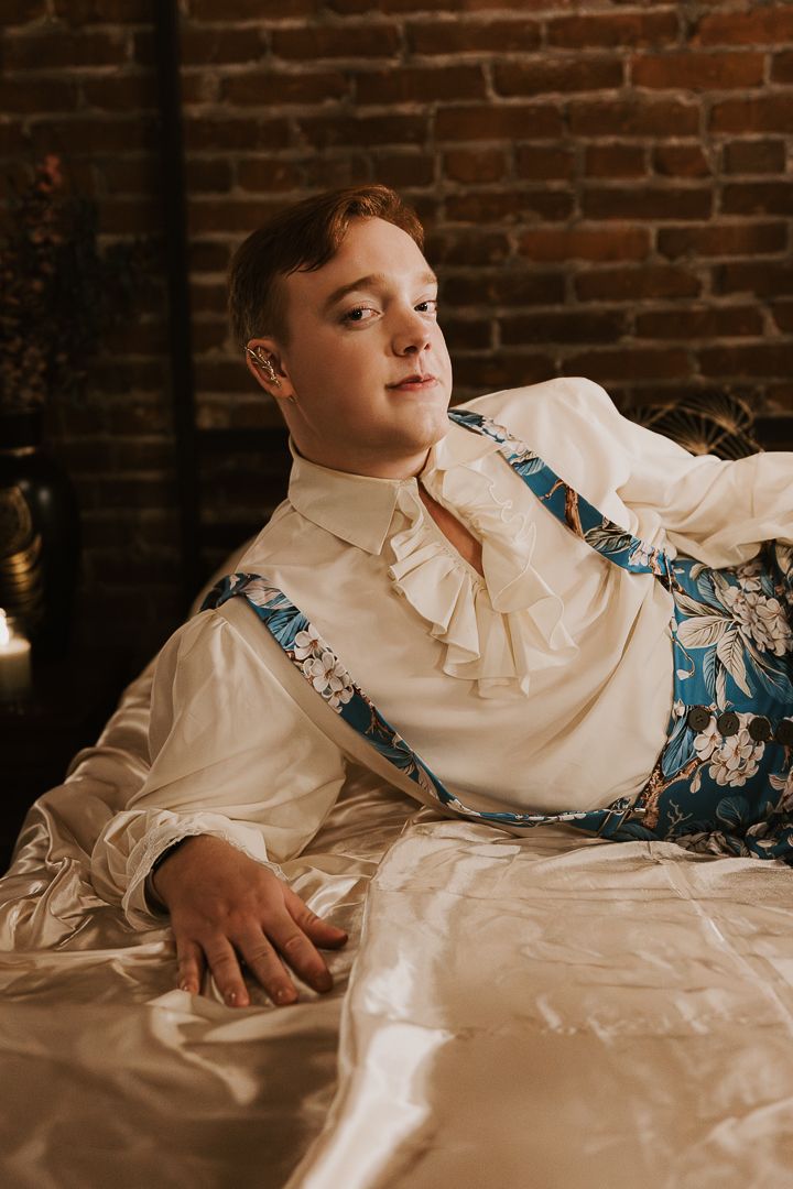 A person in a white collared shirt and floral print robe reclines on white bedding against exposed brick walls.