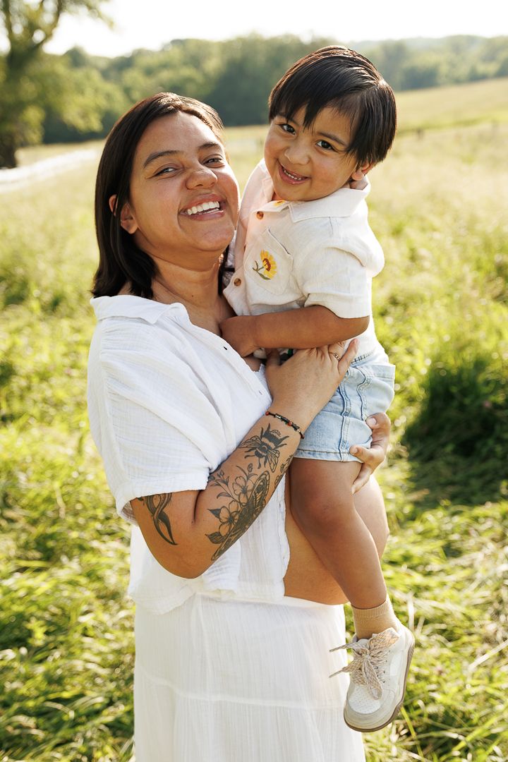 Joyful parent and child share a tender outdoor moment in a sunny meadow wearing white and denim.