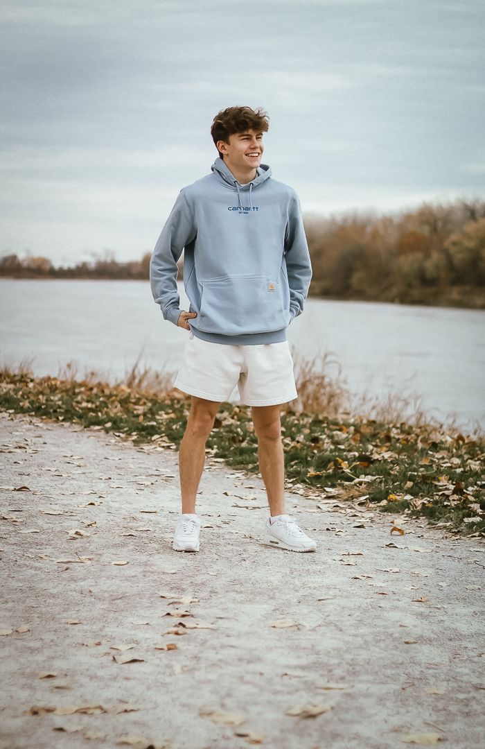 Someone in a light blue hoodie and white shorts stands on a rocky beach path during autumn with water in the background.