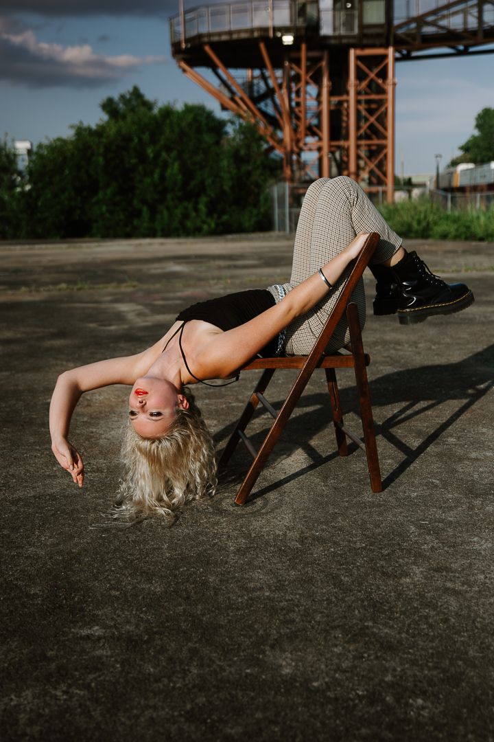 Artistic fashion shoot on industrial ground with model reclining on wooden chair against metal structure backdrop.