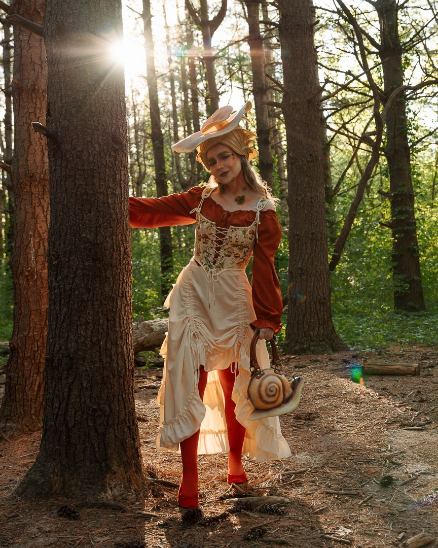 Woman in Renaissance costume with snail purse poses in sunlit forest wearing red stockings and ornate corset dress.