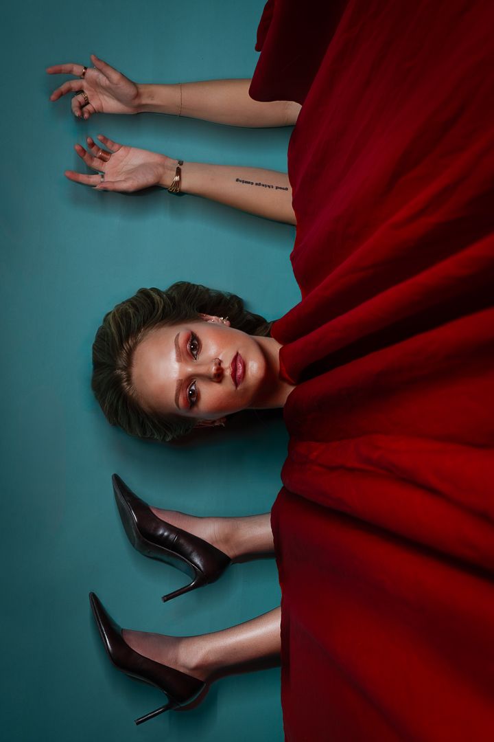 A fashion portrait against turquoise backdrop featuring red fabric and black stiletto heels in a dramatic pose.