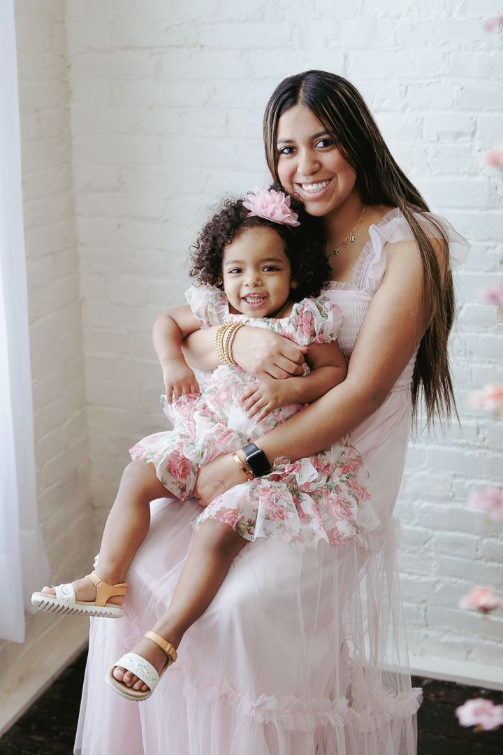 Mother and daughter in matching pink dresses embrace while posing in a bright studio with white brick walls.