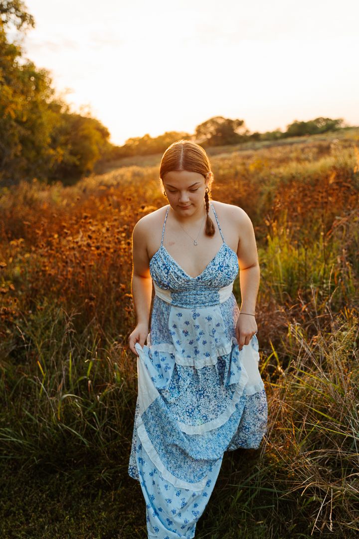 A blue floral maxi dress flows in the golden sunset light among tall grasses in a rural field.