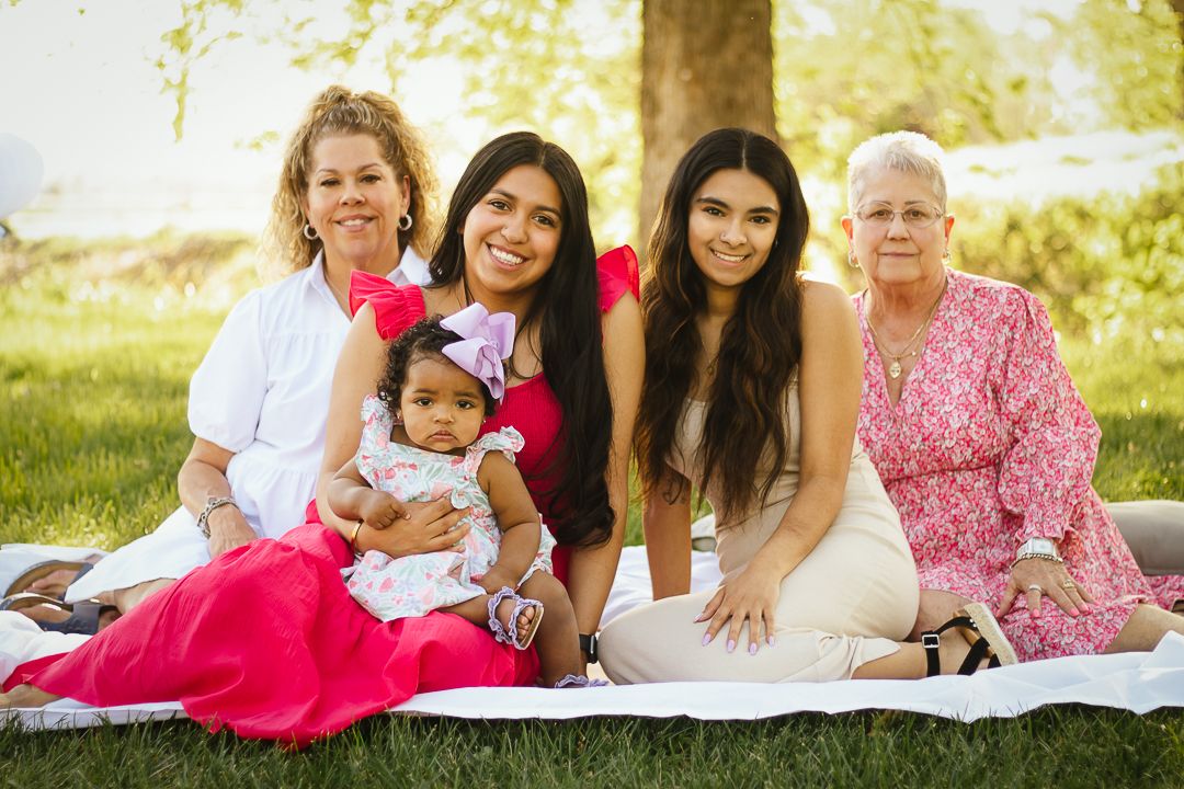 A multigenerational family lying together outdoors on a white blanket in a sunny park setting.