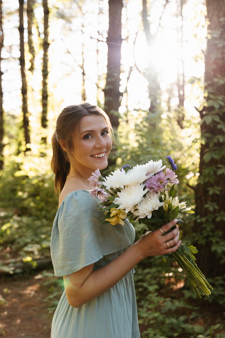 Someone wearing a mint green dress holds a bouquet of pink and white flowers in a sunlit forest setting.