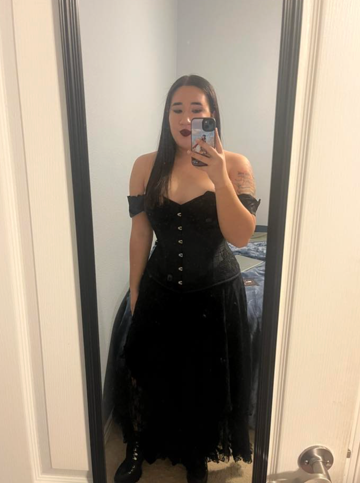 Mirror selfie showing someone wearing a black sleeveless dress and taking photos in a bathroom with white walls.