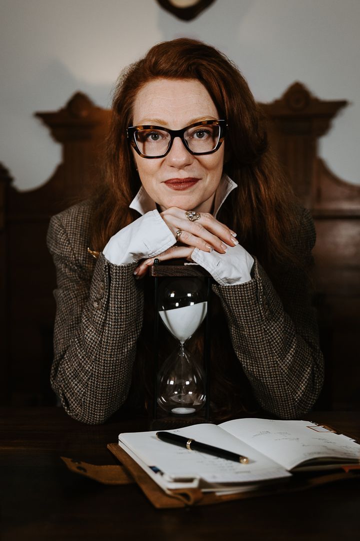 Professional wearing glasses and tweed jacket sits at desk with beverage while studying from open book in dark setting.