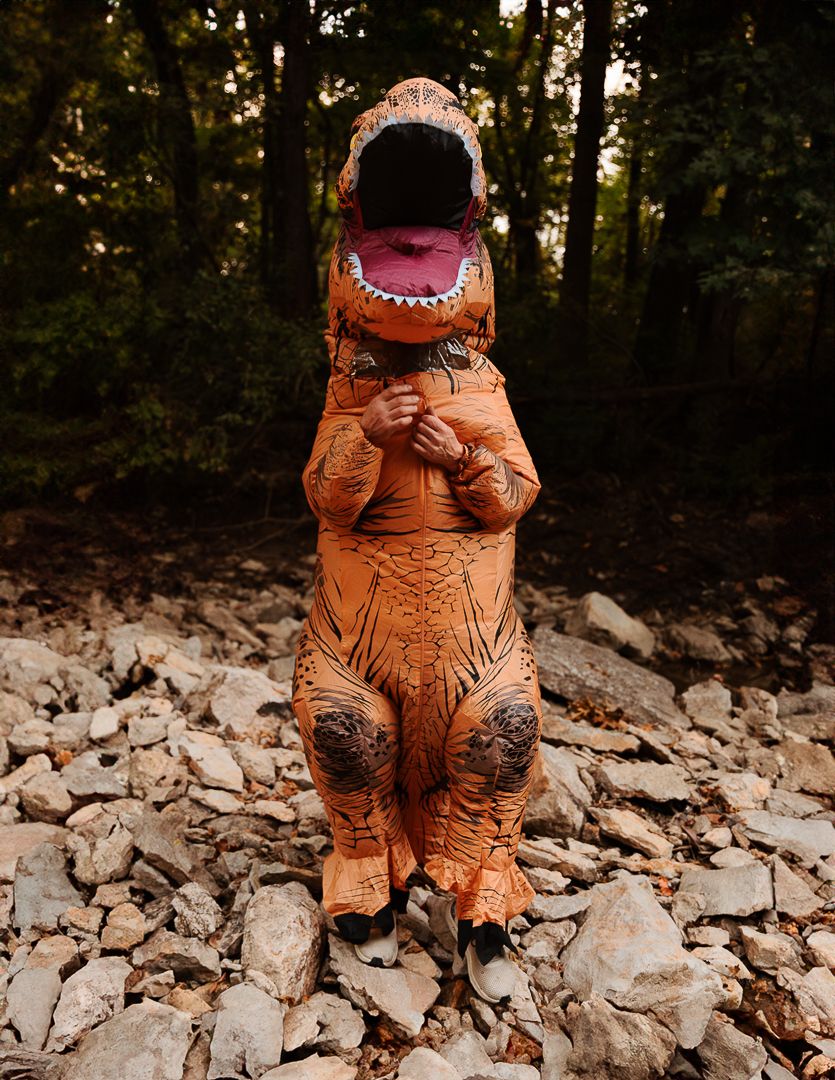 Orange inflatable T-Rex dinosaur costume photographed on rocky terrain in a forest.