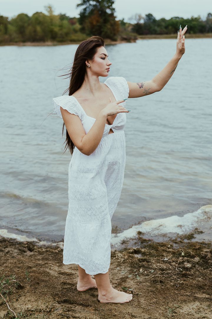 Woman in flowing white dress dances gracefully on a rocky beach shore.