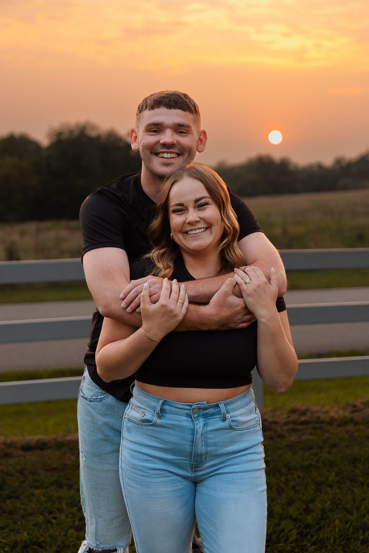 A couple embraces during sunset while standing in a grassy field, creating a silhouette against the orange sky.