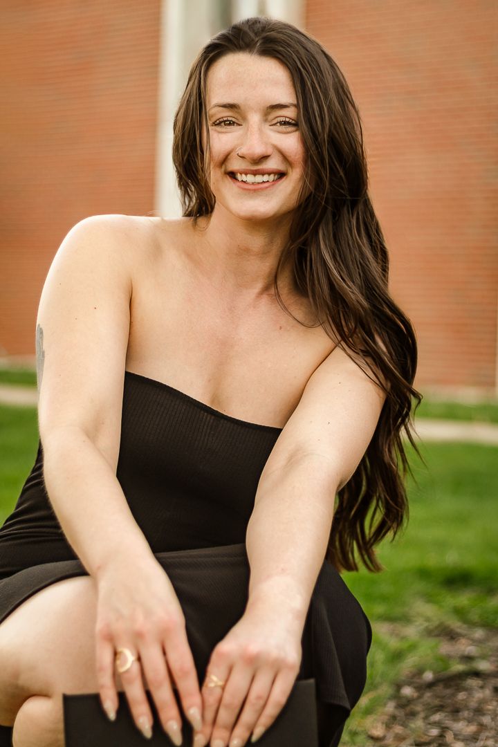 A woman in a black top smiles and poses against a brick wall with grass in the background.