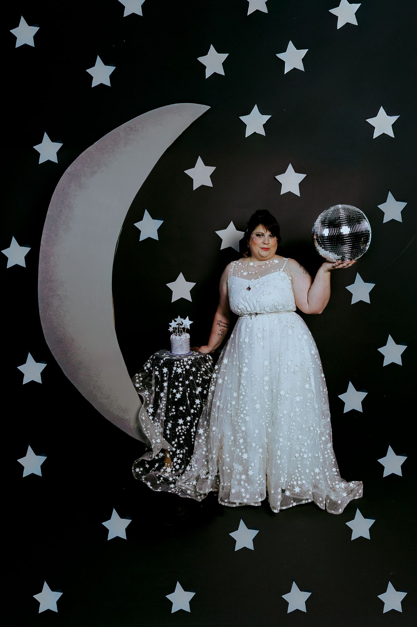 Plus-size bride in star-embroidered white gown poses with disco ball beside crescent moon on starry black backdrop.