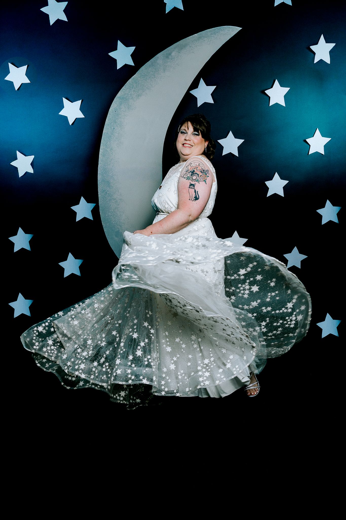 Woman in star-patterned white gown poses on crescent moon prop with star backdrop.