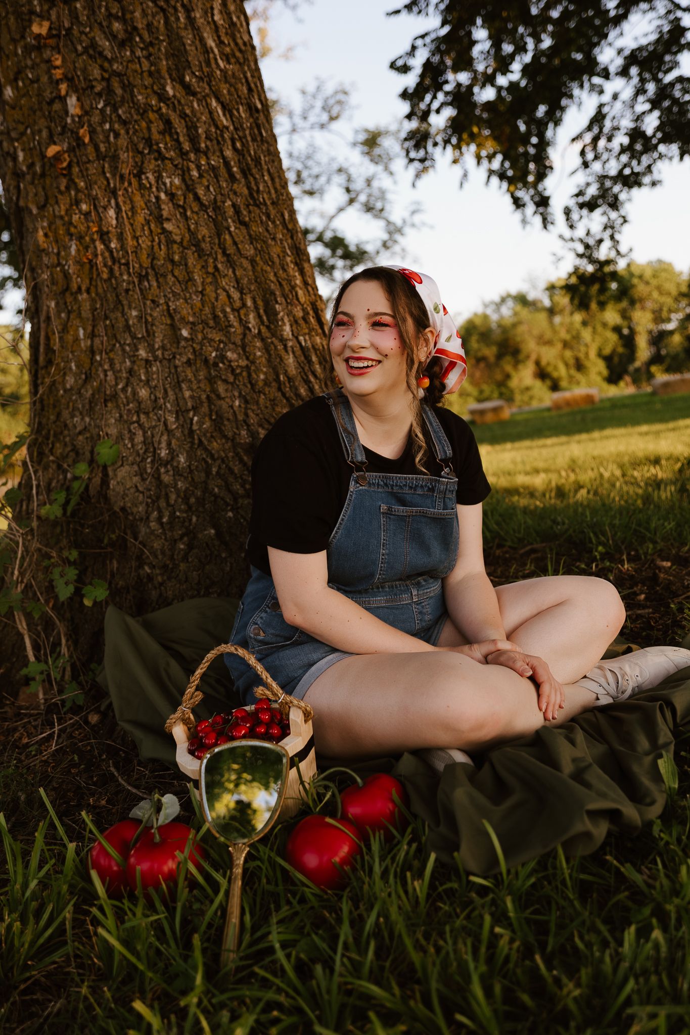 Young woman in denim overalls sits under a tree with a basket of strawberries and apples on a sunny day.