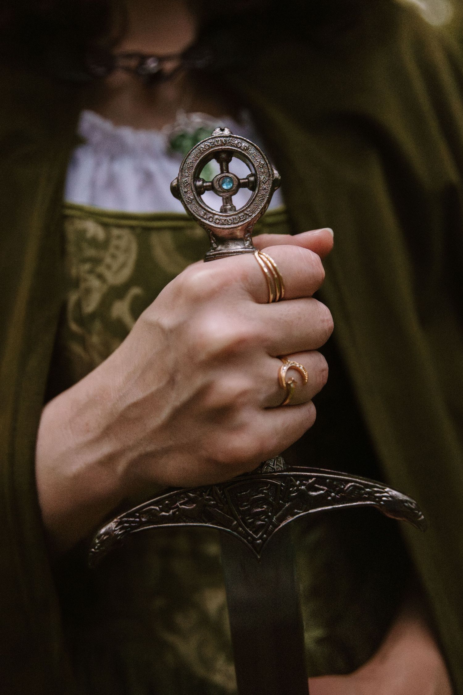 Close-up of a hand wearing gold rings gripping an ornate medieval sword hilt with Celtic cross design.