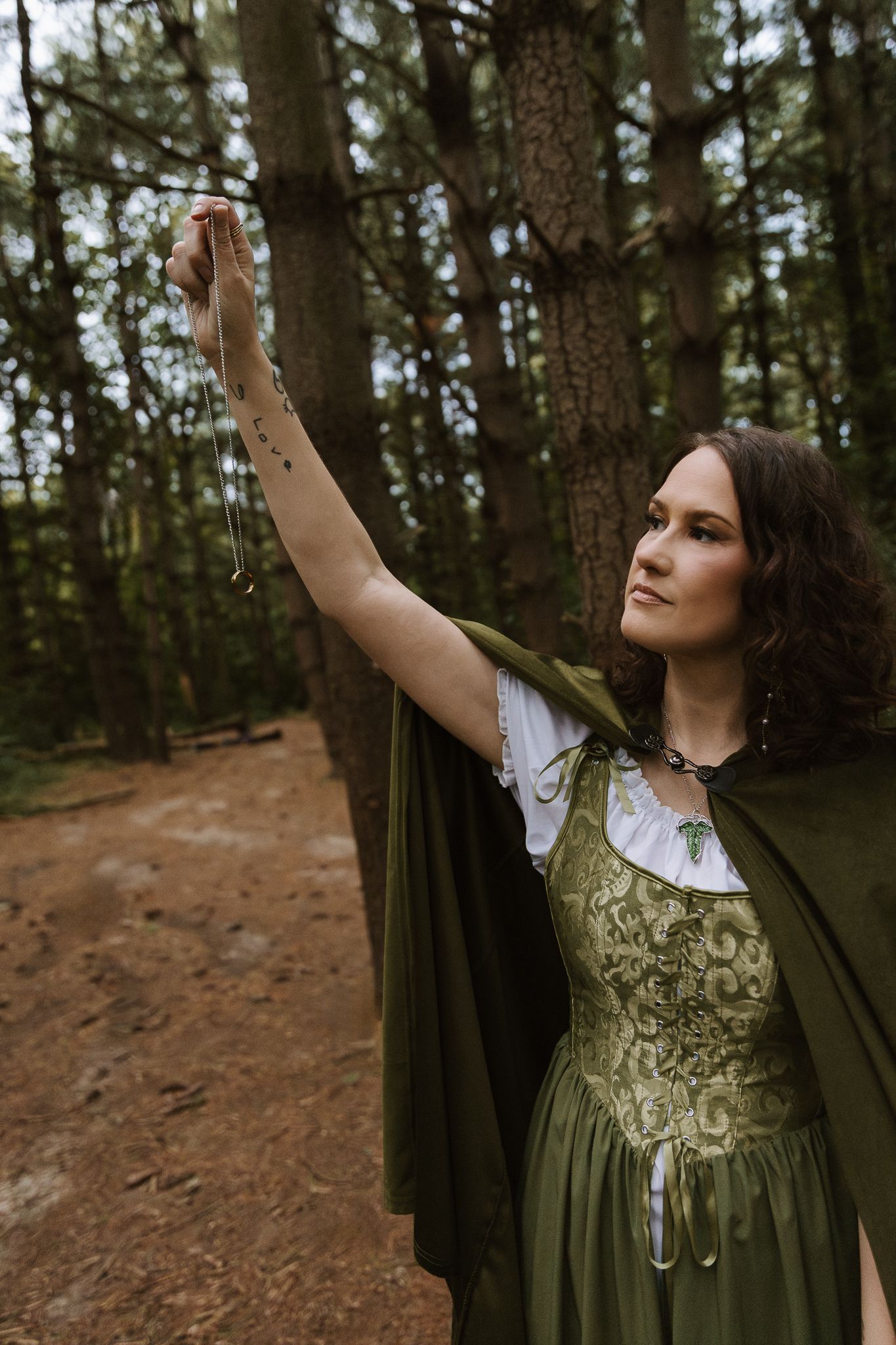 Woman in green medieval dress and cape holding a necklace in a pine forest.