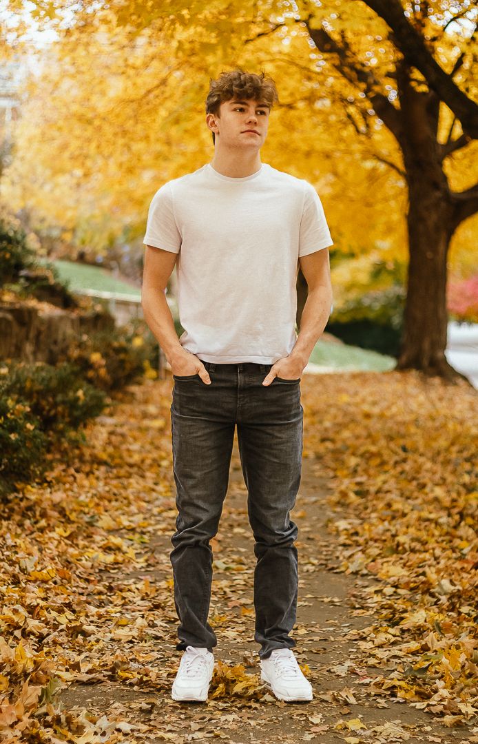 Person in white t-shirt and dark jeans stands on fall path covered in golden leaves beneath autumn trees.
