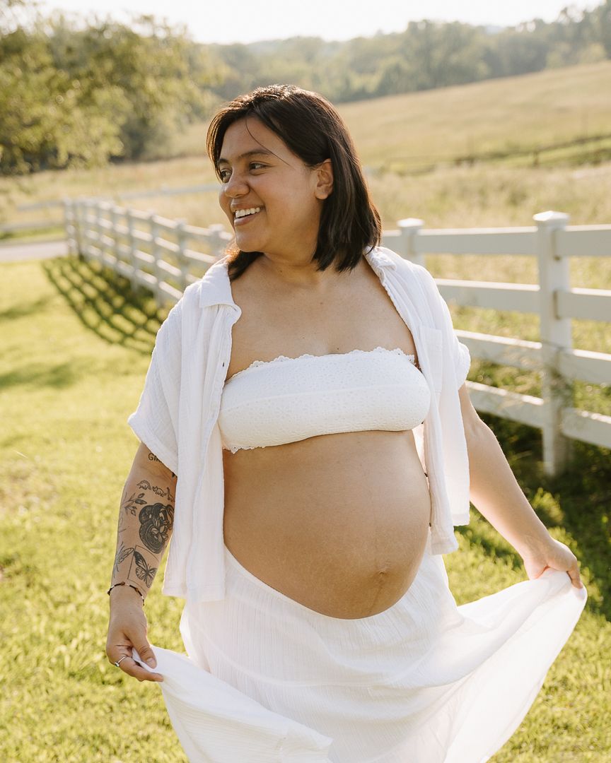 Maternity photo series in a sun-drenched field with white fencing, featuring a flowing white dress against golden light.