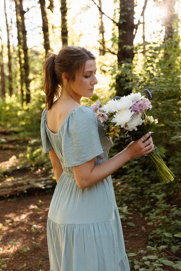 A person in a light blue dress holds a bouquet of white and purple flowers in a sunlit forest setting.