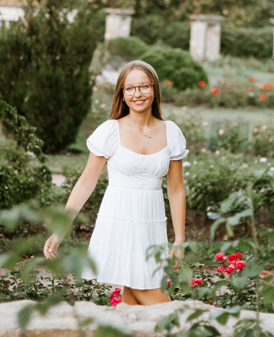 White dress-clad woman poses gracefully among blooming flowers in a peaceful garden setting.