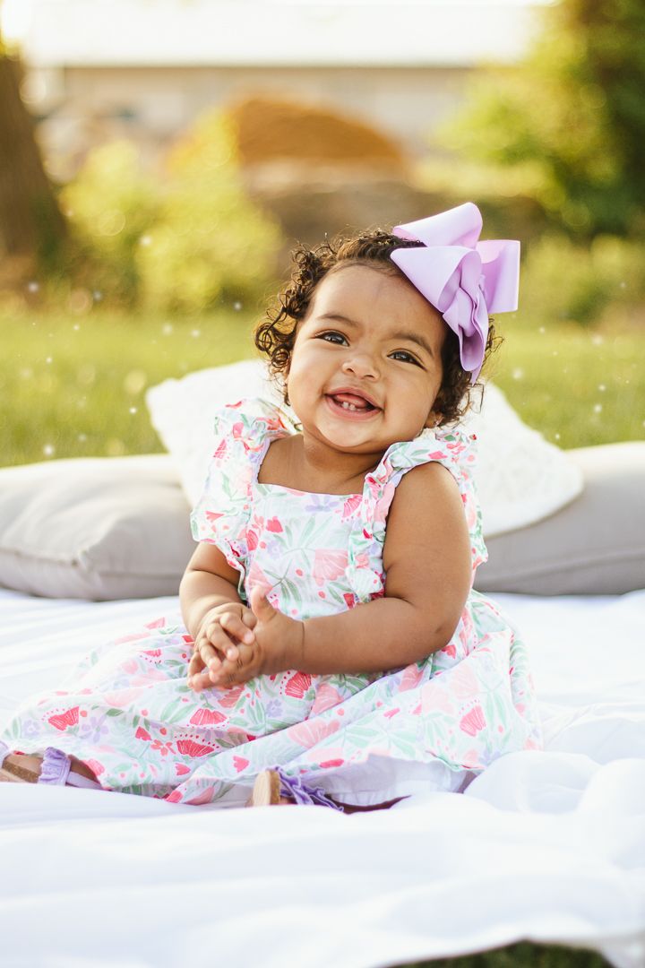 A baby in a floral dress lies on white bedding while playing outdoors on a sunny day.