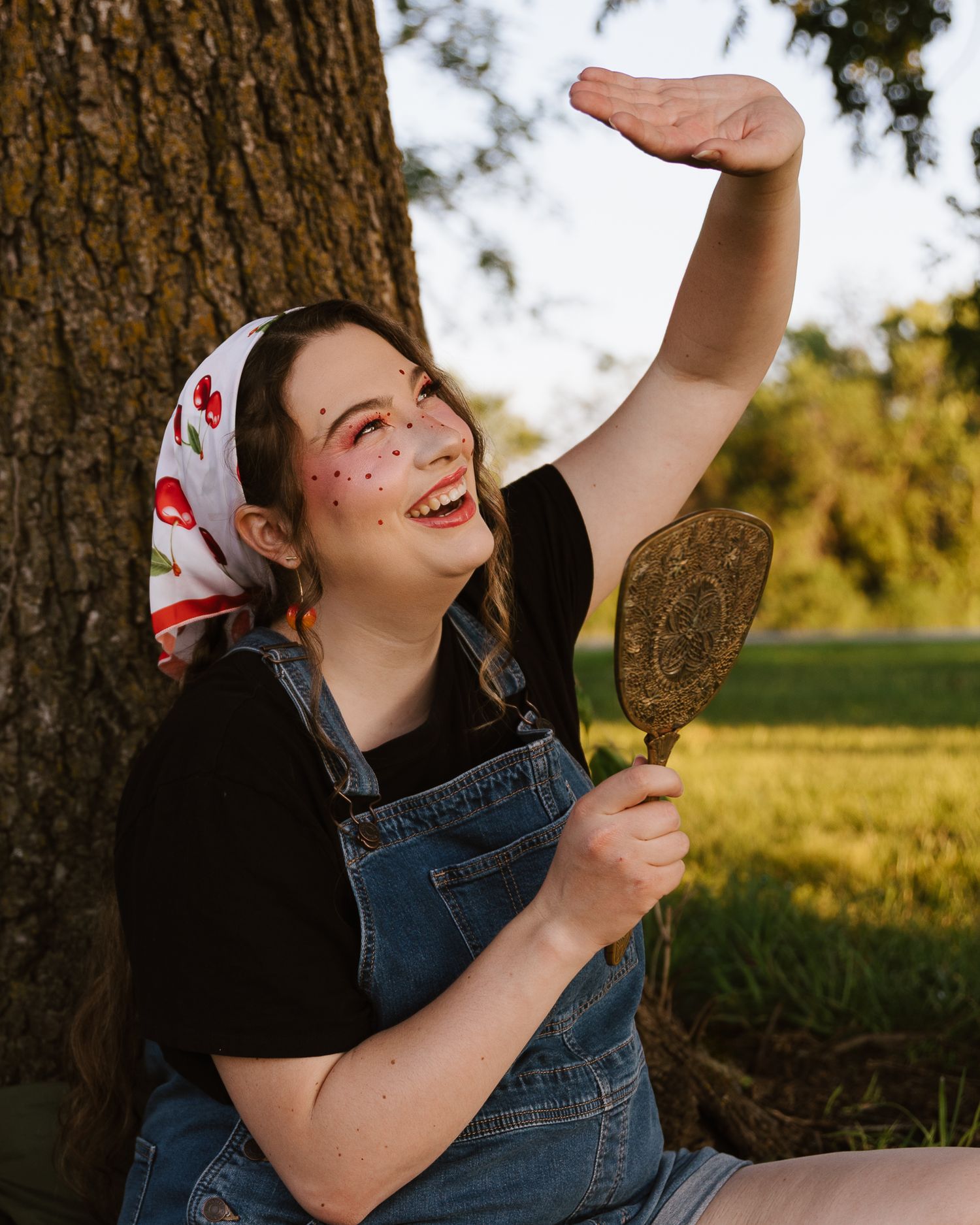 Young woman in denim overalls and cherry bandana holding vintage mirror while sitting against tree outdoors.