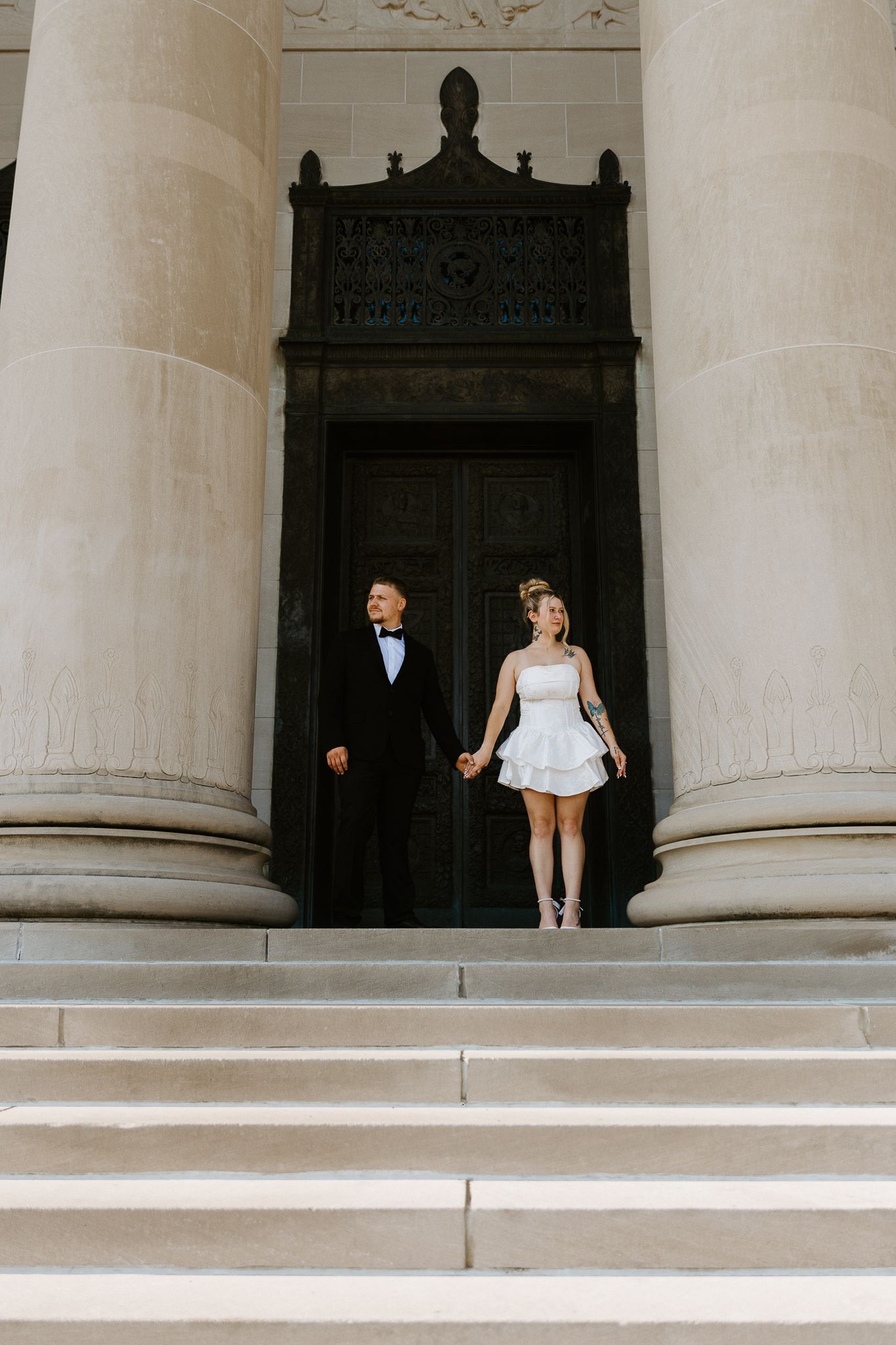 Bride in short white dress and groom in black tuxedo holding hands on grand stone steps before ornate doors.