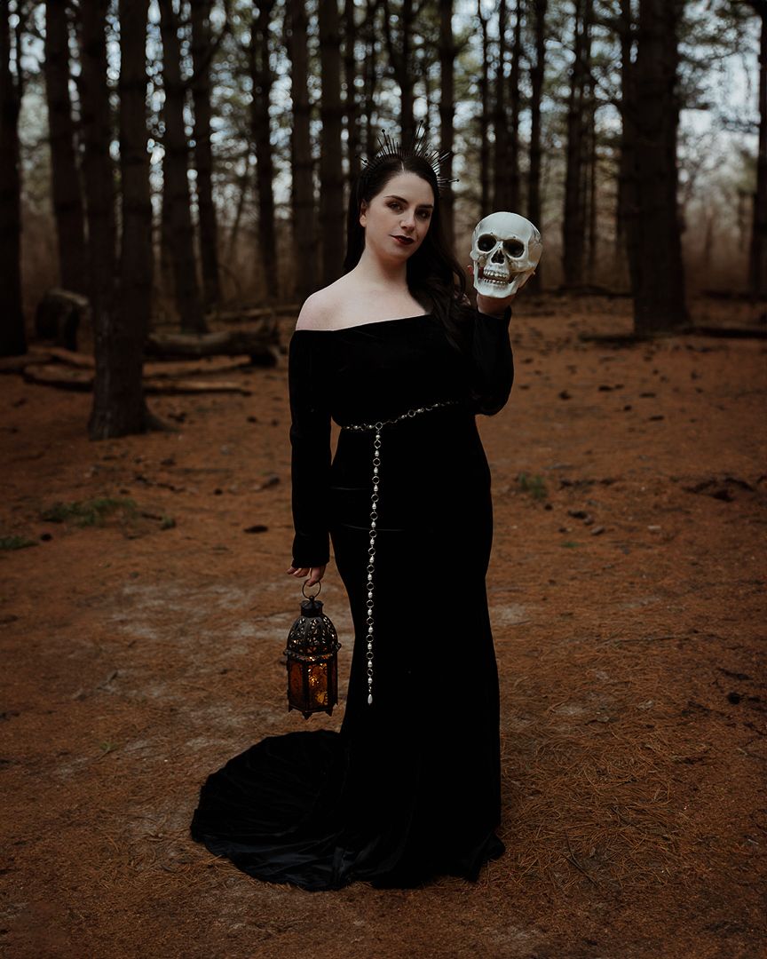 Woman in black off-shoulder gown holds skull and lantern in misty forest, gothic Halloween portrait.