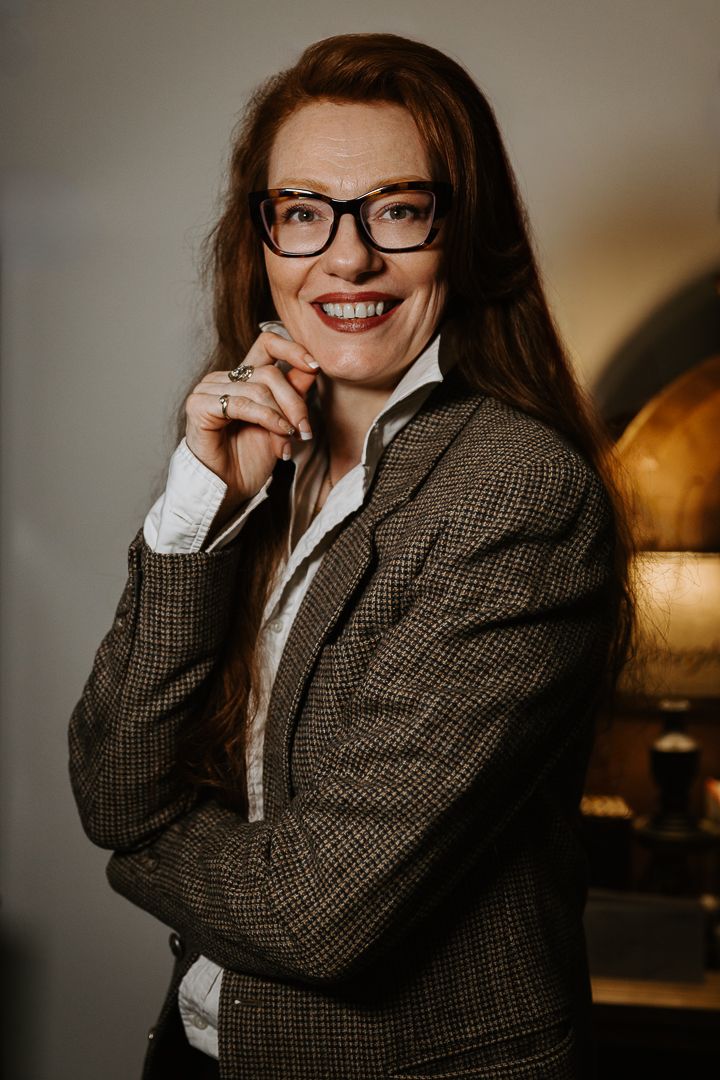 Professional portrait of someone in a brown tweed blazer and glasses smiling warmly with hand near their face.