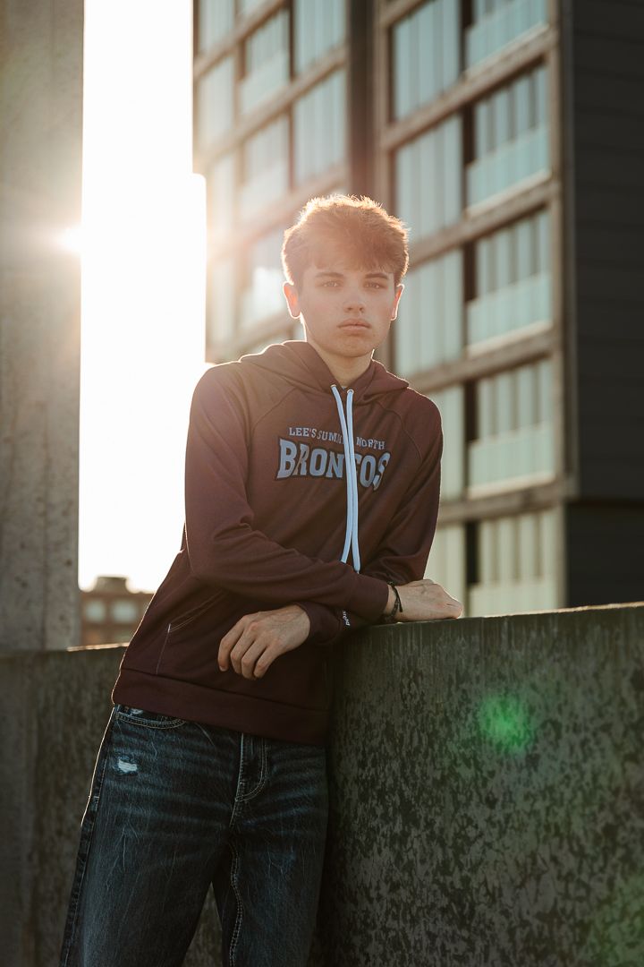 A moody backlit portrait of someone in a dark hoodie leaning against an urban building at sunset.