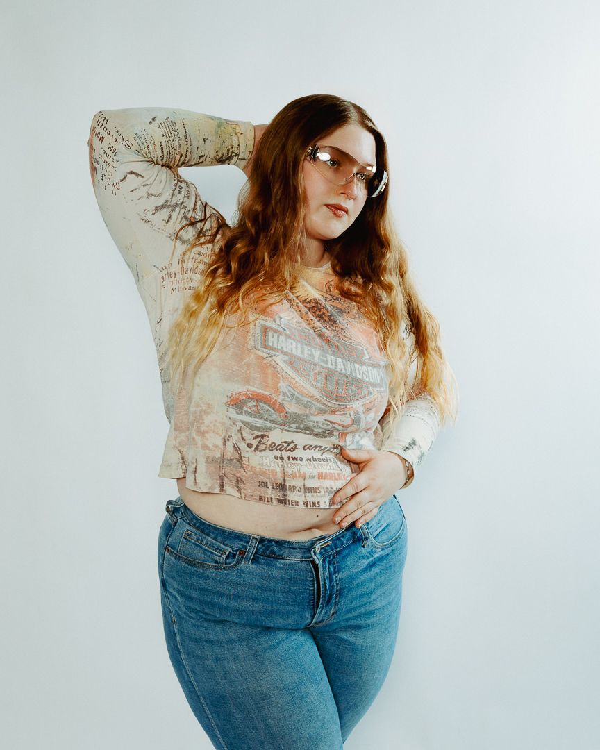 Person wearing a cream colored sweater and blue jeans poses against white wall in fashion photography series.