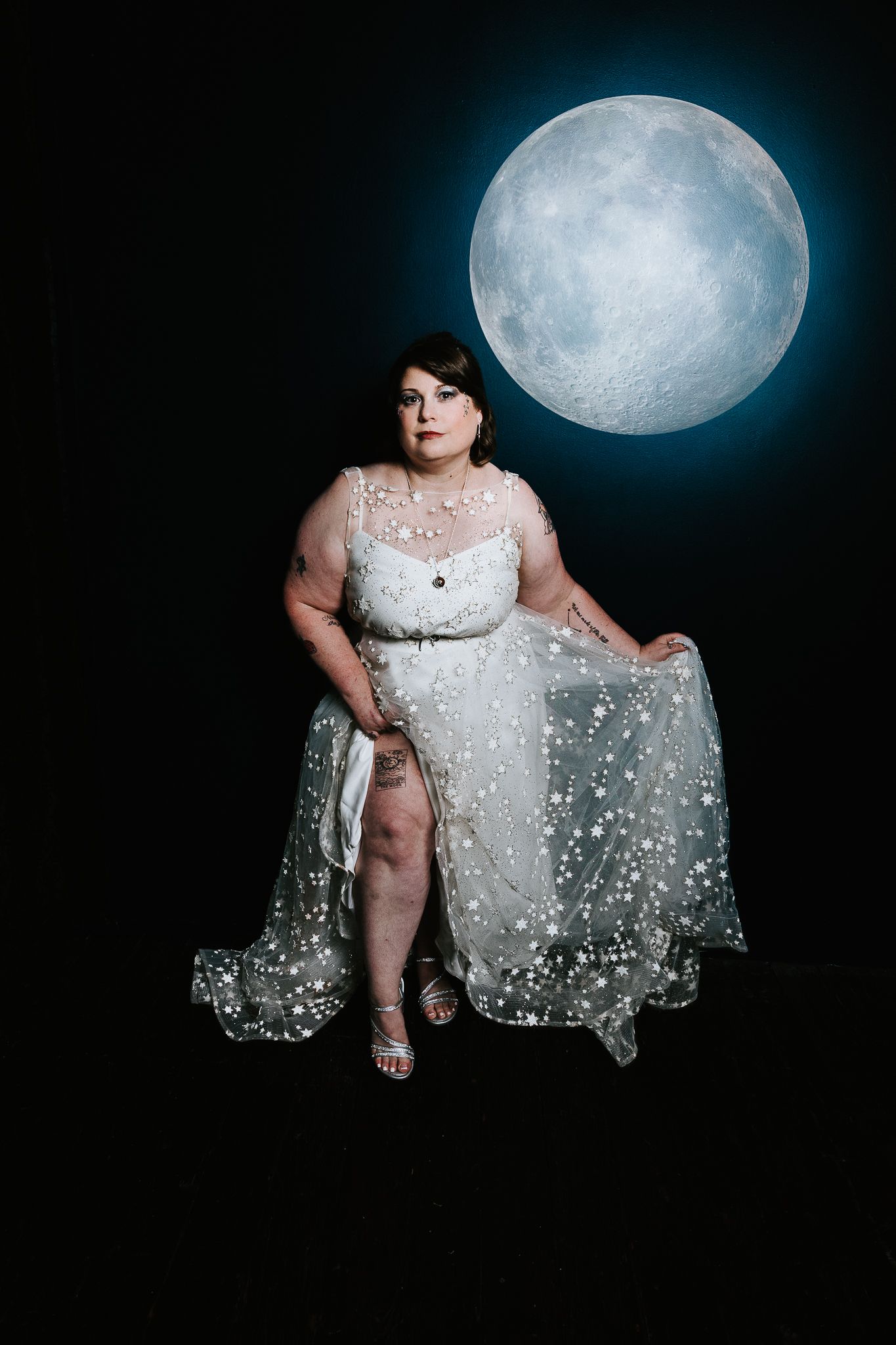 Plus-size bride in star-embellished white gown poses elegantly before a glowing full moon backdrop.