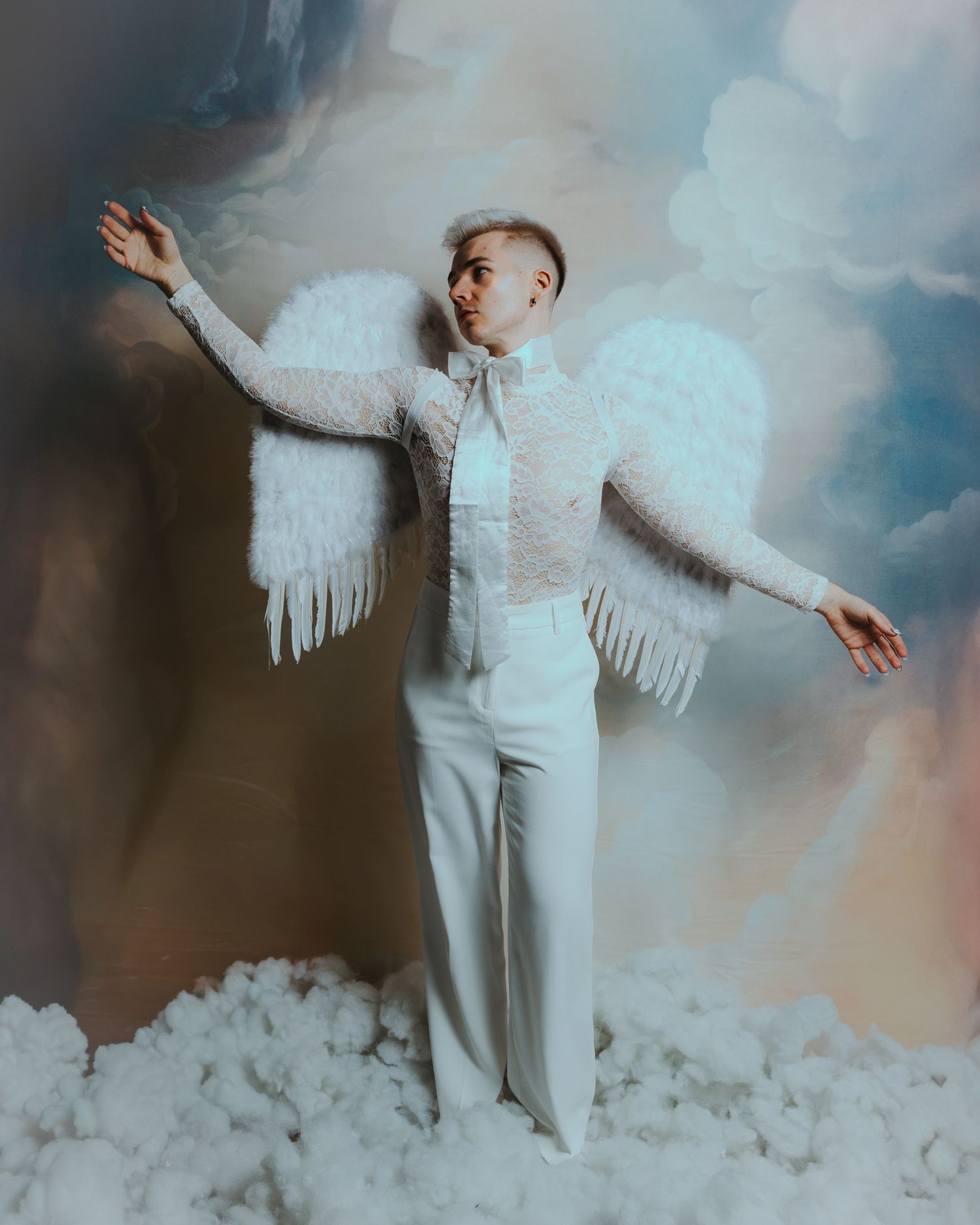 Person dressed as an angel in white lace outfit with feathered wings, posing dramatically against a cloudy sky backdrop.