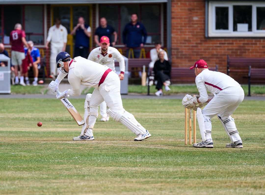 Grassroots cricket action as the batsman defends a delivery while wicketkeeper and slip fielder prepare for a catch.