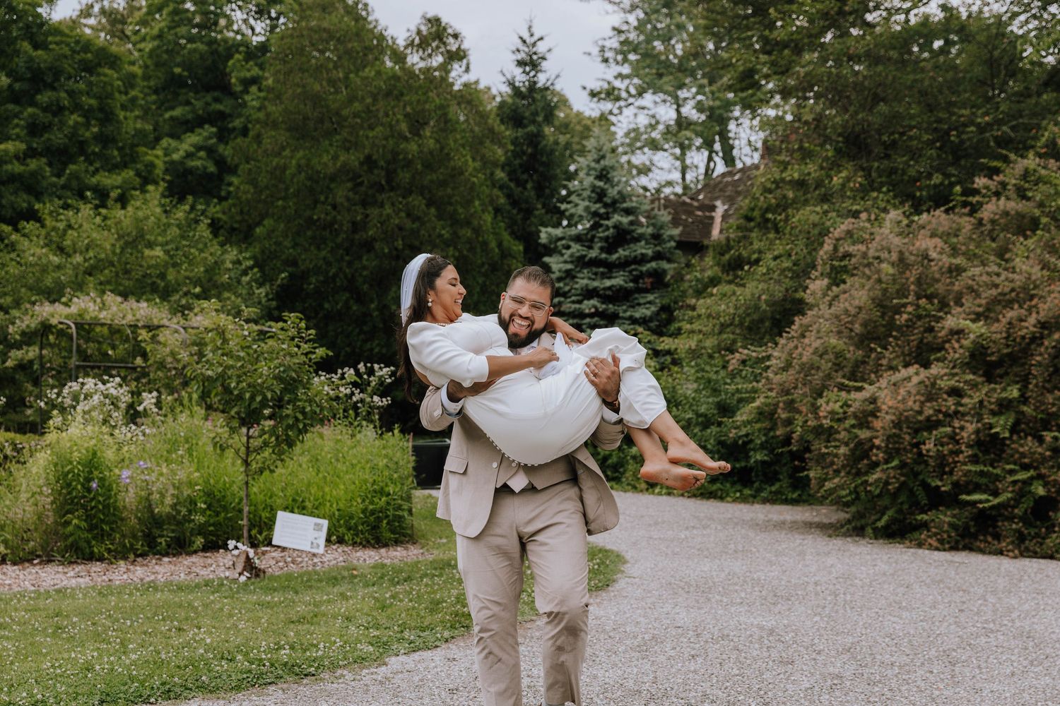 Groom laughing as he victoriously carries his shoeless bride across a rock path