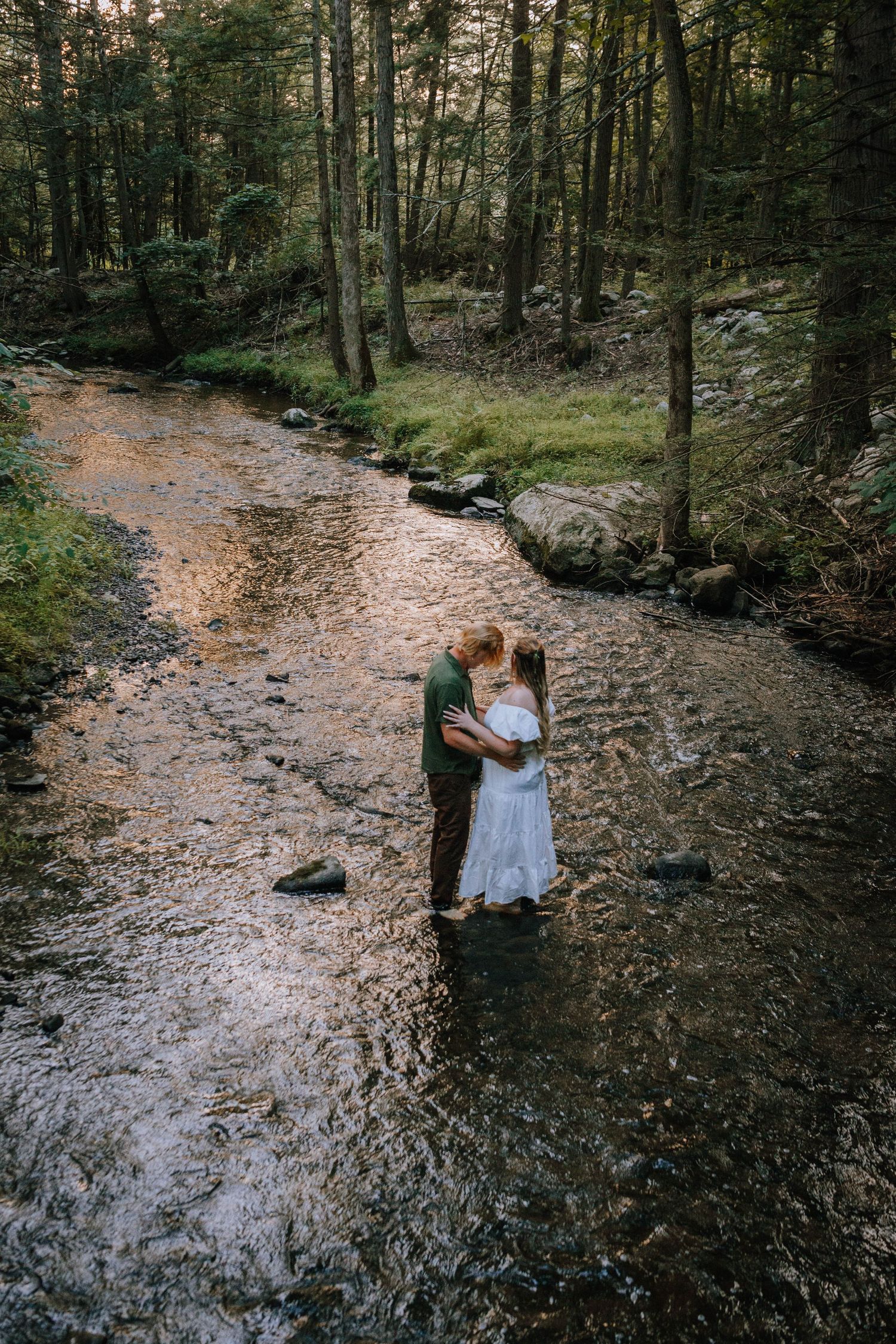 an engaged couple hold each other while balancing on a rock in the middle of a creek reflecting the sunset