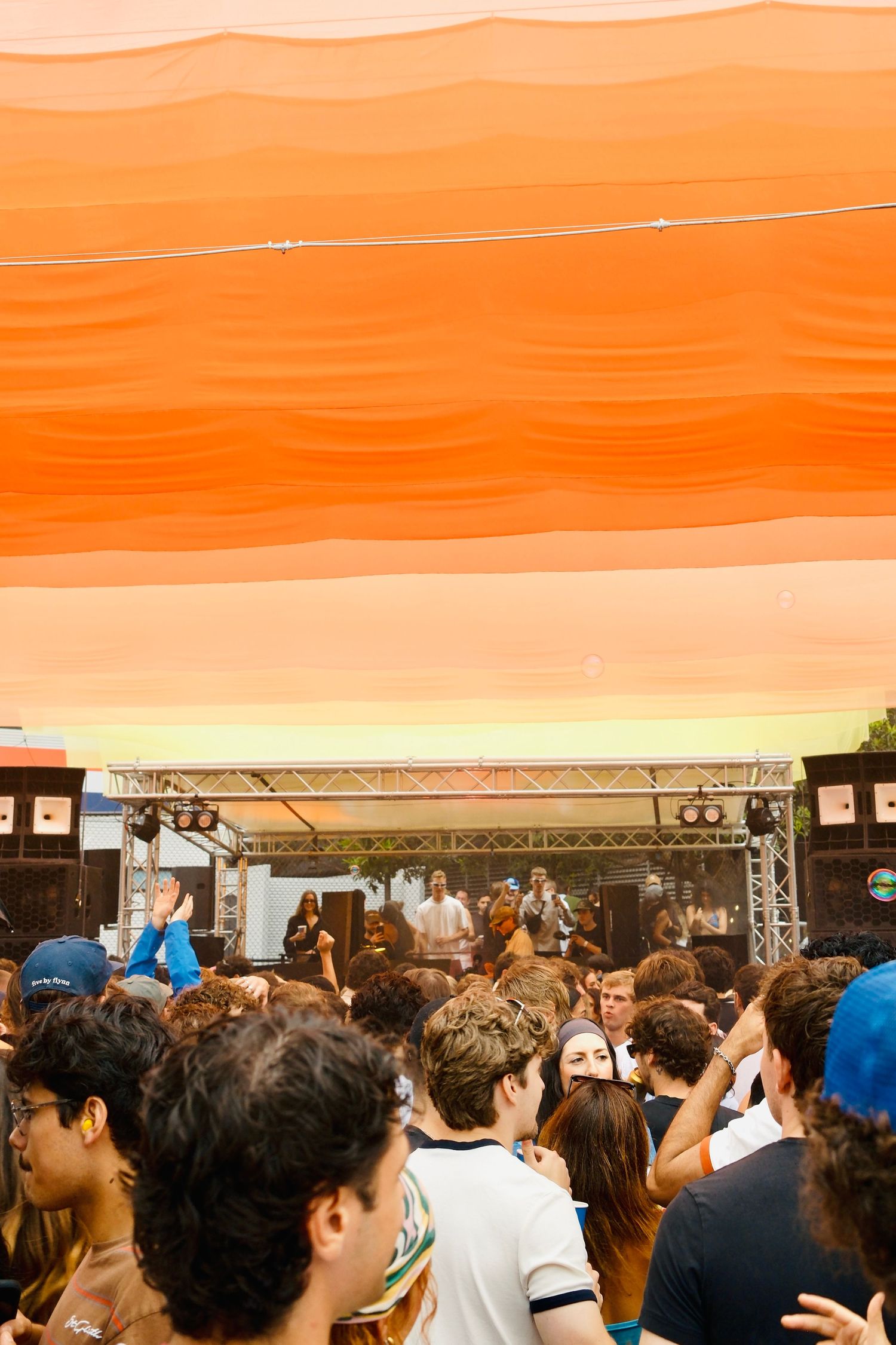 Large crowd at outdoor music festival with orange canopy and band performing on stage.