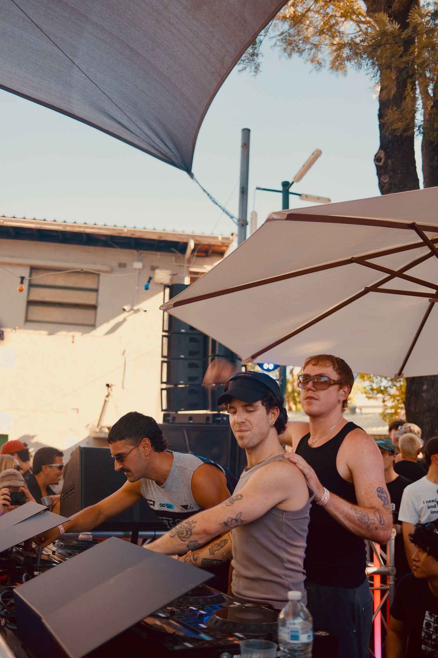 Three tattooed DJs performing at an outdoor daytime music festival under a shade canopy and umbrella.