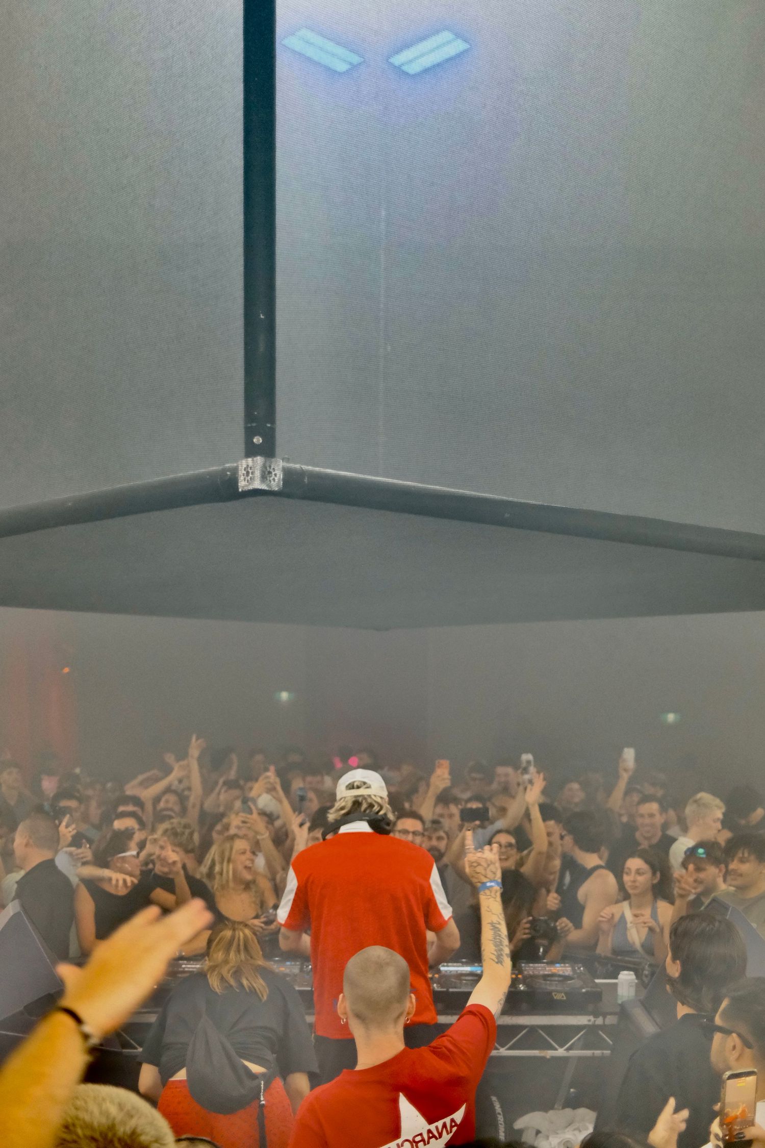 DJ in red outfit performing on stage before a large fog-filled crowd at an indoor music venue.